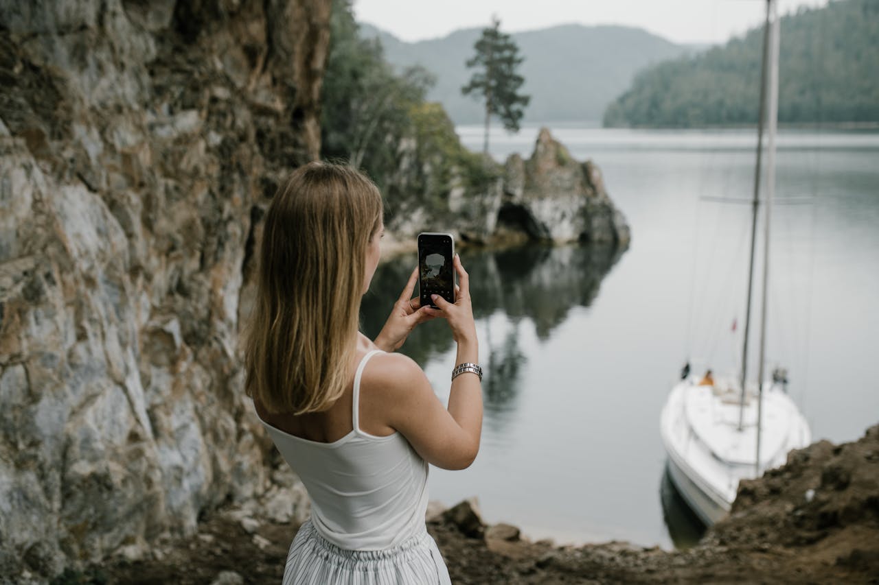 A woman takes a photo of a serene lakeside with a sailboat in lush natural surroundings.